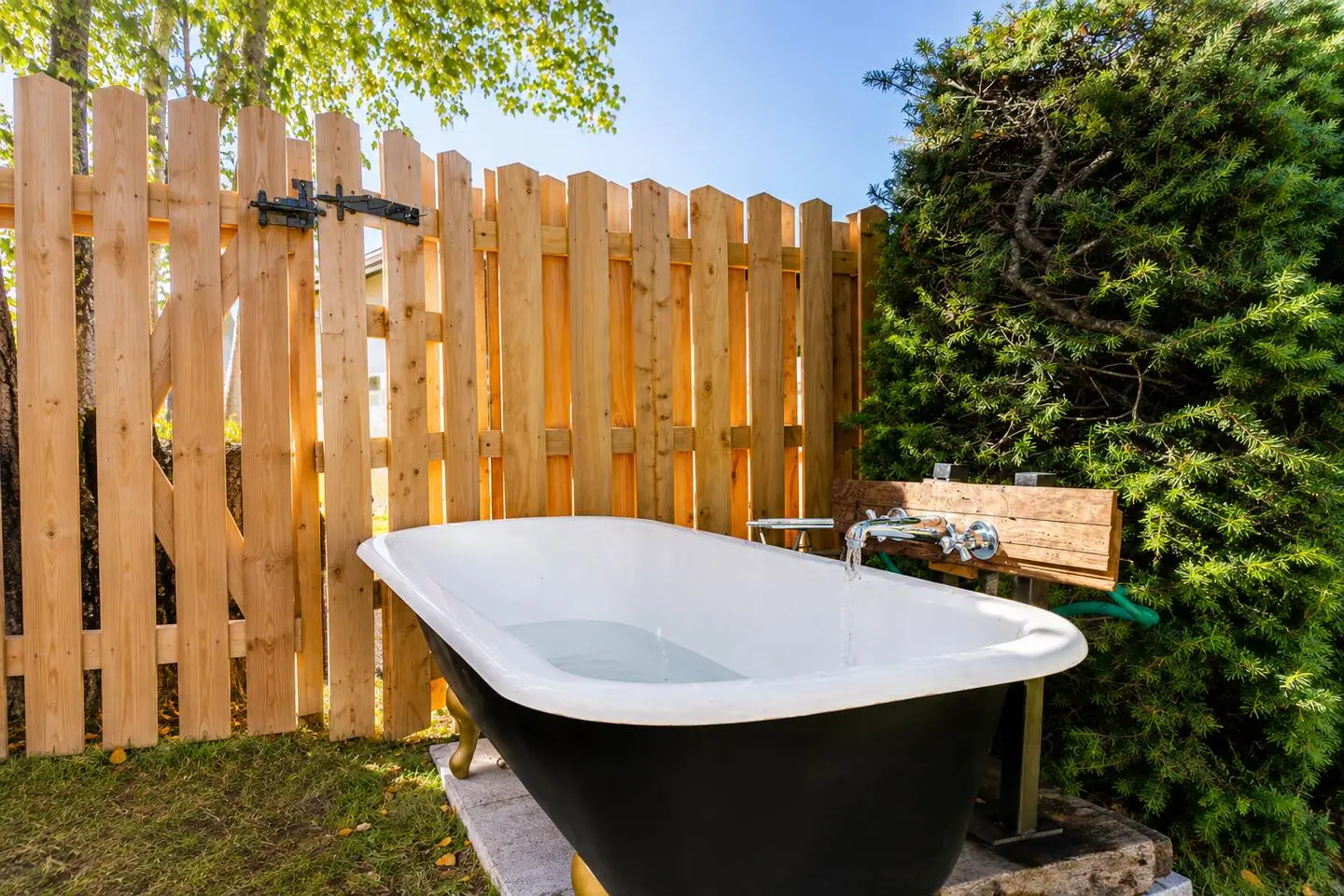 Outdoor clawfoot bathtub filling with water, set against a wooden fence and lush greenery.