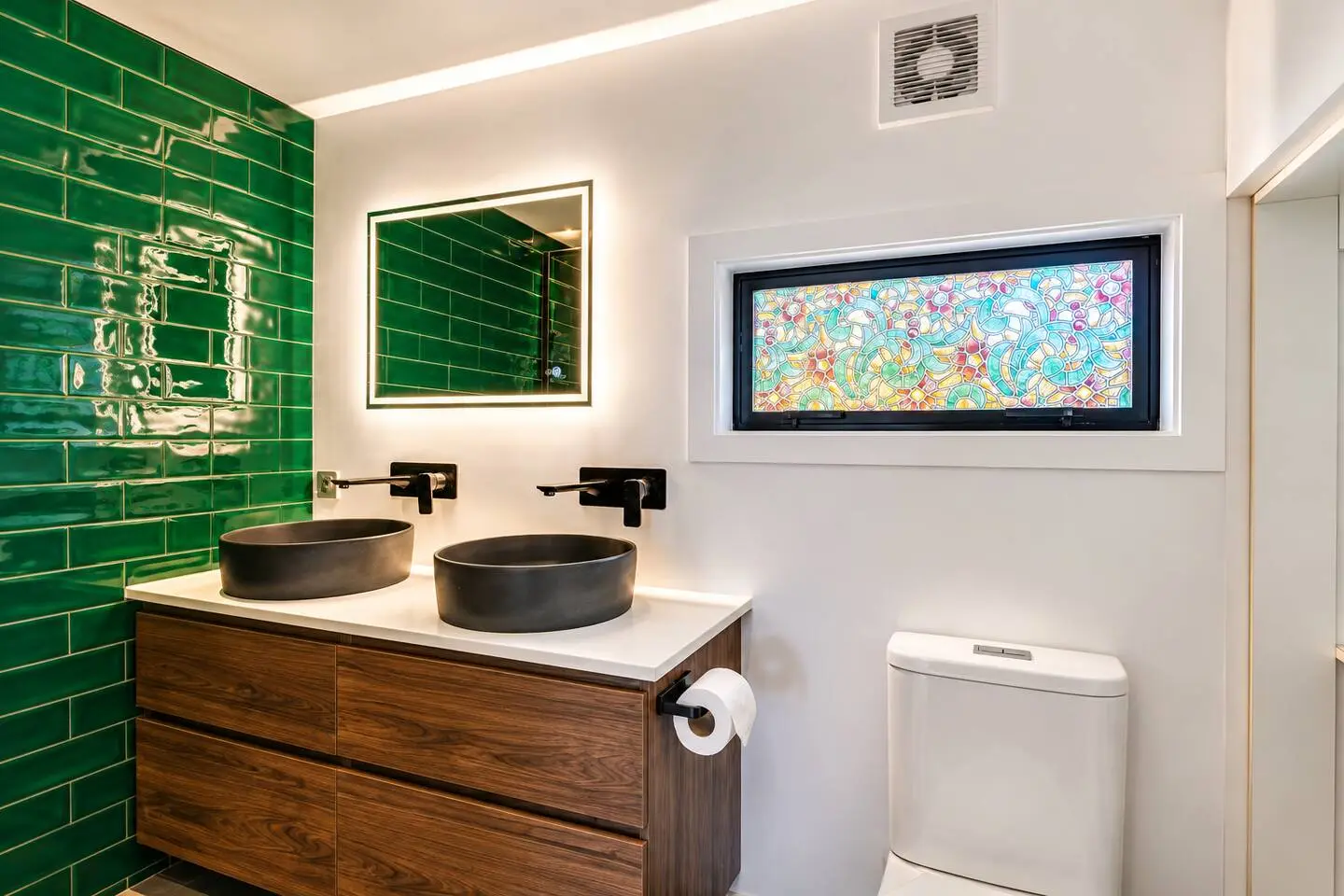 Modern bathroom with green subway tile, dual vessel sinks, walnut vanity, and a colorful stained-glass window.