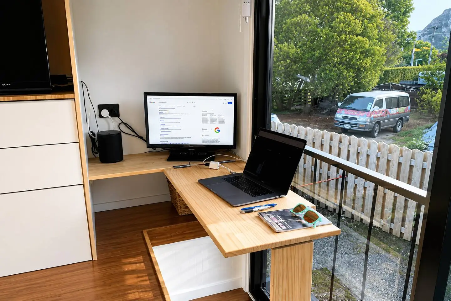 Wooden desk workspace with monitor, laptop, and speaker, beside a window overlooking a fenced driveway.