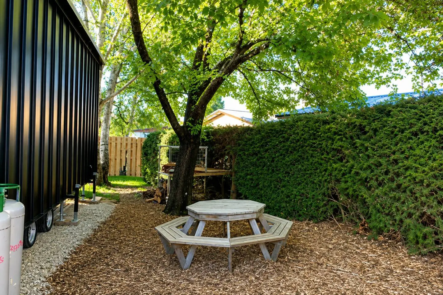 Octagonal picnic table under a shady tree beside the tiny house, with tall hedges surrounding the yard.