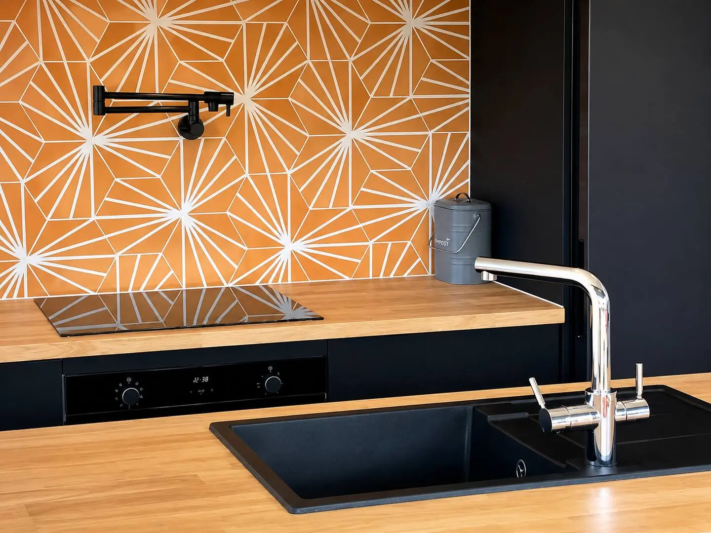 Close-up of kitchen sink, faucet, and induction stove against the geometric orange tile backsplash.